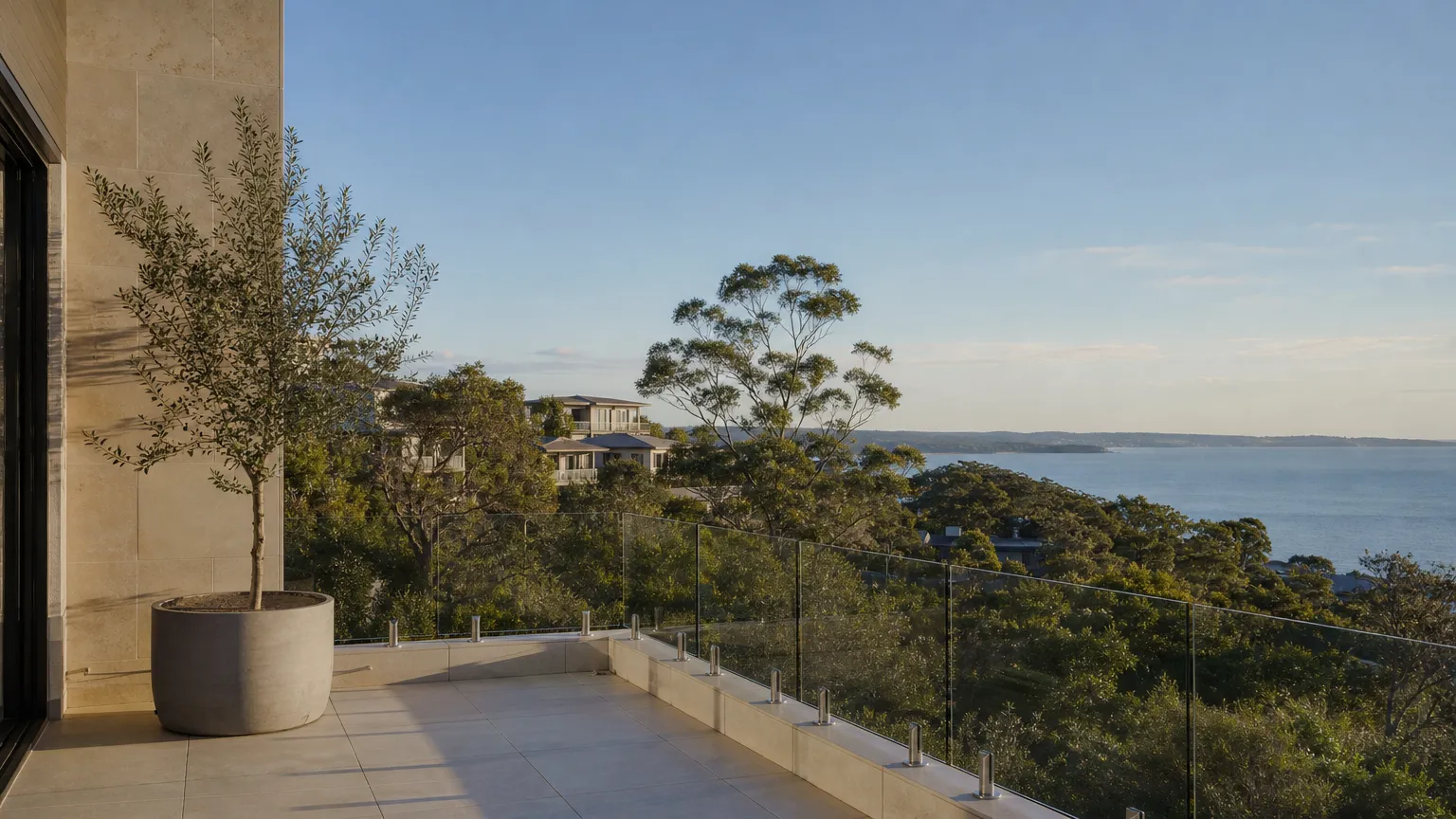 a frameless glass balustrade on stainless spigots along the tiled upstand of a balcony, looking over gum trees to a wide bay