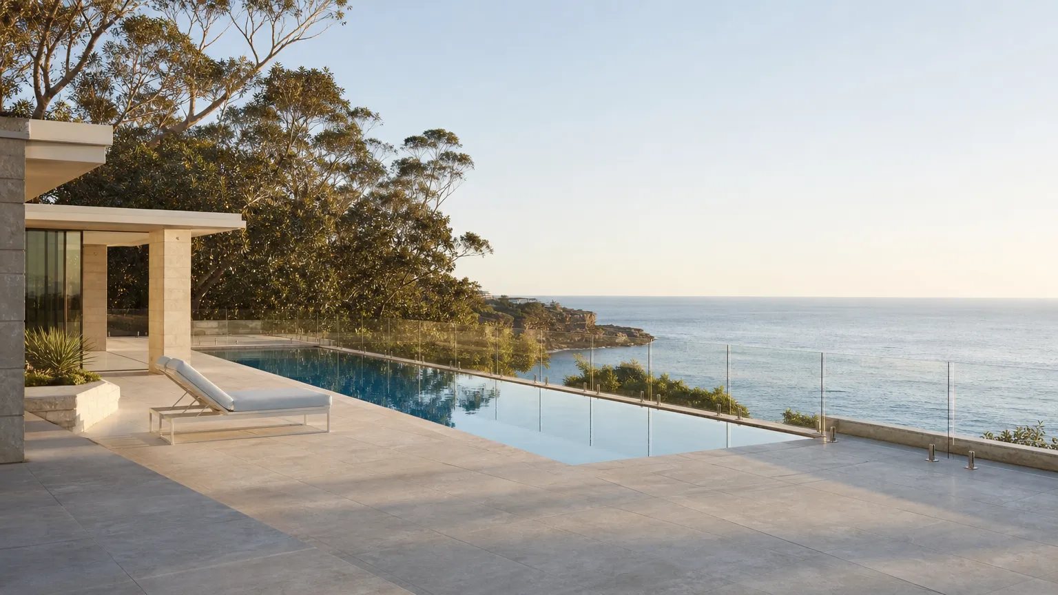 a frameless glass pool fence on stainless spigots along the edge of a clifftop infinity pool, with the ocean and a sandstone headland behind