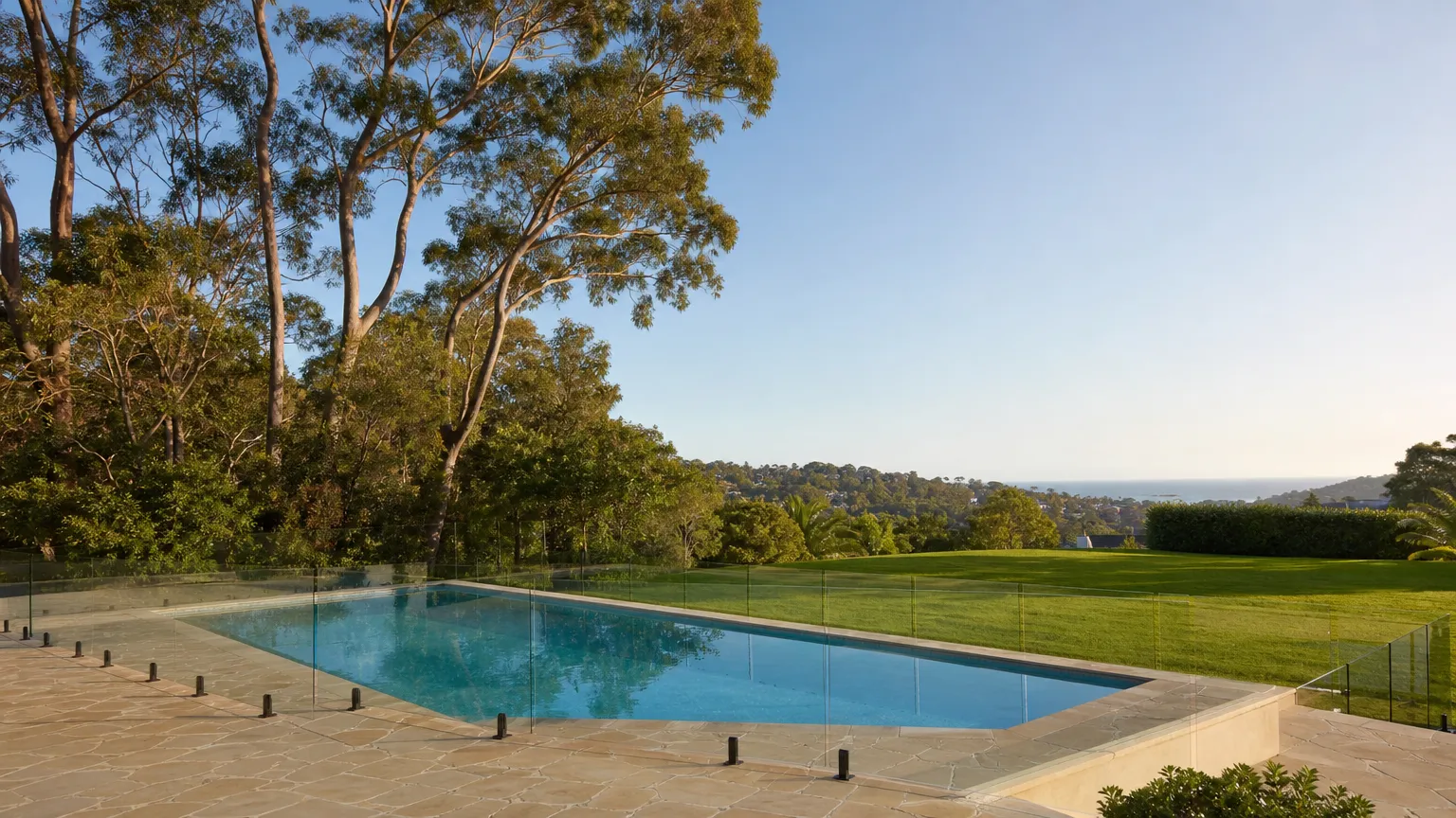 a semi-frameless glass pool fence running along a wide lawn with large gum trees behind